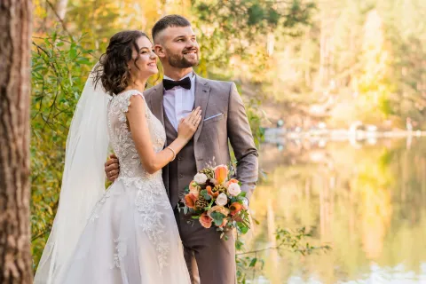 Bride and groom in wedding attire embracing near a serene lake surrounded by autumn trees and soft sunlight.