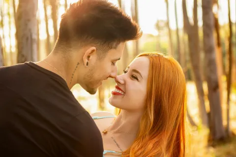 Young couple embracing and smiling at each other in a sunlit forest during golden hour.