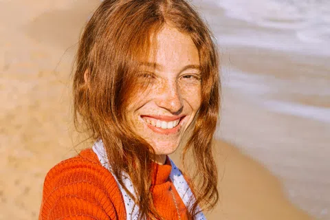 Smiling red-haired woman with freckles wearing an orange sweater on a sunny beach.