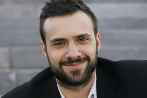 Close-up portrait of a smiling man with beard and dark hair wearing a black blazer sitting outdoors on steps.