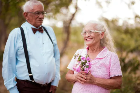 Elderly couple outdoors smiling, woman holding pink flowers, man wearing suspenders and bow tie in soft natural setting.