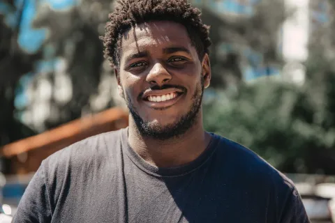 Smiling young man in a dark t-shirt outdoors with blurred trees and buildings in the background