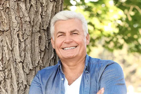Smiling senior man in a denim shirt leaning against a tree, exuding warmth and confidence in natural light.