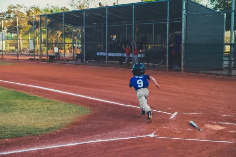 Young baseball player in blue jersey running from home plate toward first base on a sunny day
