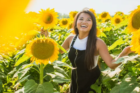 Young woman smiling in a vibrant sunflower field on a sunny day wearing a white shirt and black dress.