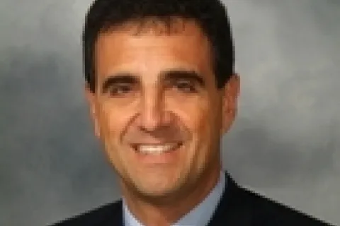 Smiling man in a suit with a red tie, set against a gray background.