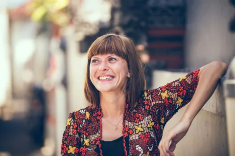 Woman in colorful floral blouse smiling and leaning against a wall outdoors in soft sunlight.