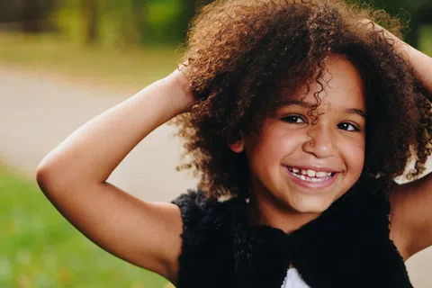 Smiling young girl with curly hair and missing front teeth wearing a black vest outdoors in soft natural light