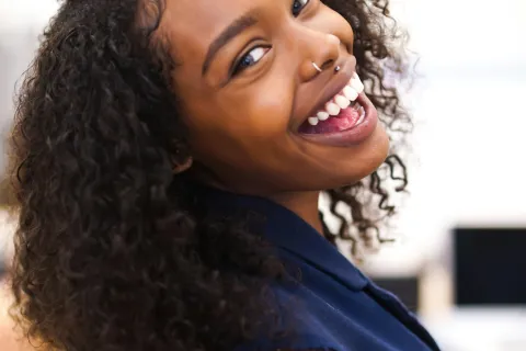 Smiling woman with curly hair and nose ring wearing a sleeveless navy top in a bright room