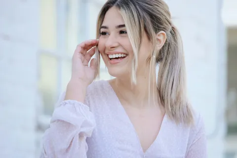Smiling young woman with blonde hair in ponytail wearing light purple blouse outdoors
