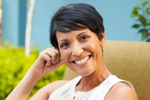 Smiling woman with short hair sitting outdoors, enjoying a sunny garden setting.