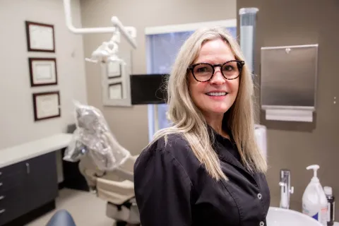 Smiling female dentist in black coat standing in a modern dental clinic with equipment and certificates.