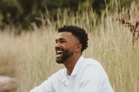 Smiling man in white shirt and shorts sitting on rocks surrounded by tall grass in outdoor natural setting