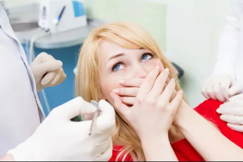Nervous woman covering mouth at dentist office surrounded by dental tools and professionals.