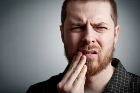 Man with beard grimacing and holding his jaw in pain against a gray background
