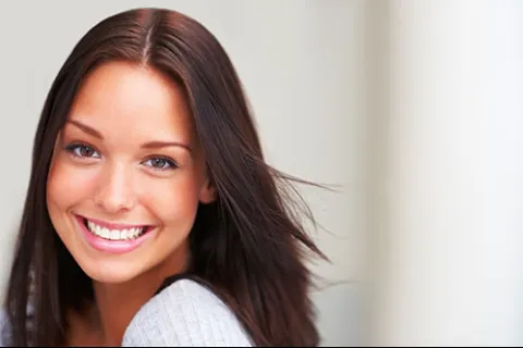 Smiling young woman with long brown hair and bright eyes wearing a light gray sweater on a neutral background