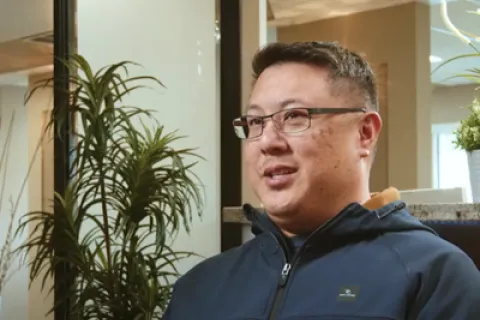Man with glasses wearing a navy blue zip-up jacket sitting indoors near a green potted plant.