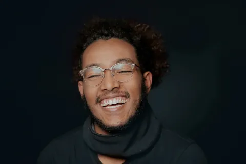 Young man with curly hair and glasses laughing against a dark background, wearing a black turtleneck.