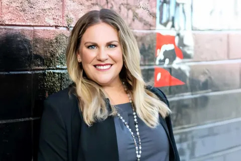Smiling woman with blonde hair wearing a black blazer and necklace standing by a graffiti wall.