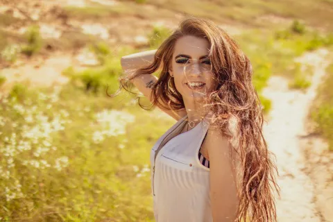 Smiling young woman with long flowing hair posing outdoors on a sunlit grassy path.