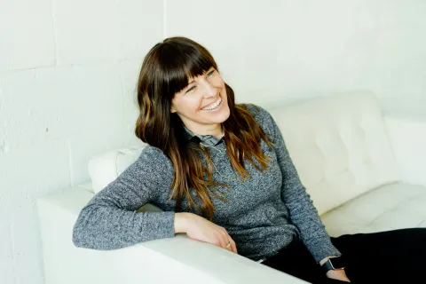 Smiling woman with long brown hair in gray sweater relaxing on white sofa against a white wall