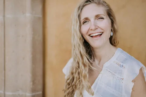 Smiling woman with long wavy hair wearing a white blouse against a warm textured wall.