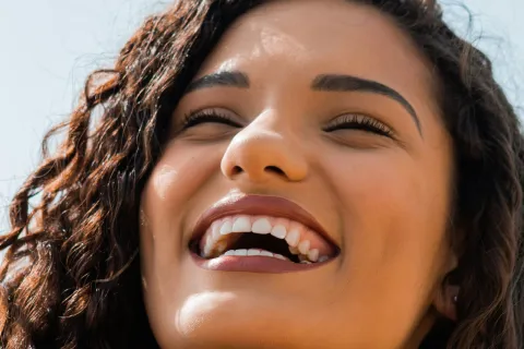 Close-up of a joyful woman with curly hair laughing against a clear blue sky, showing bright smile and red nails.