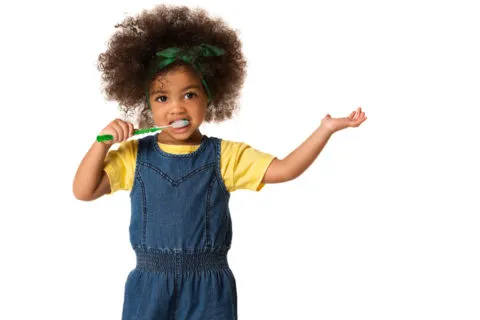Child with curly hair demonstrates brushing teeth, wearing a yellow shirt and overalls.