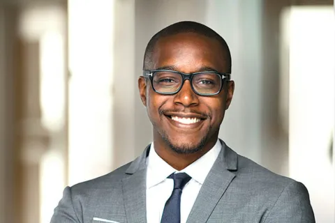 Smiling professional man in a suit with glasses, arms crossed, in a modern office setting.
