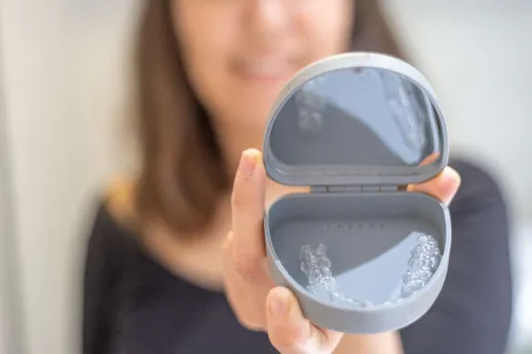Person holding a gray container showcasing transparent aligners inside, blurred background.