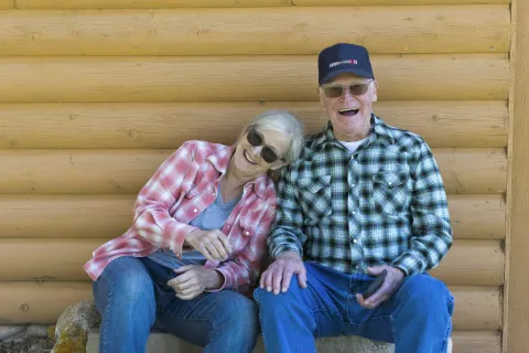 Elderly couple in plaid shirts sitting and smiling against a wooden log cabin wall on a sunny day