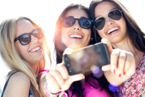Three smiling young women wearing sunglasses taking a selfie outdoors on a sunny day