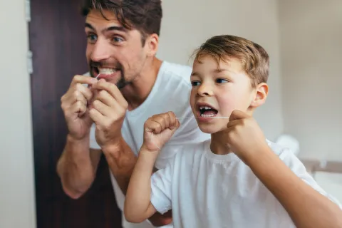 A father and son happily flossing together in a bright bathroom setting.