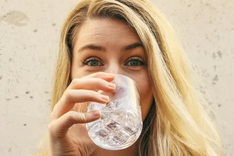 Young woman with long blonde hair drinking water from a clear glass with ice cubes against a plain background.
