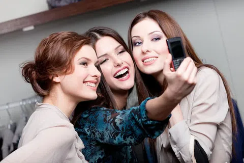 Three young women smiling and taking a selfie together using an old-style flip phone indoors.