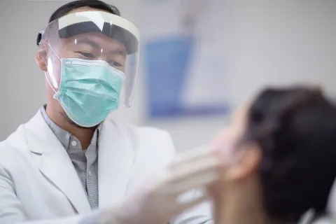 Dentist wearing a face shield and mask attending to a patient in a clinical setting.