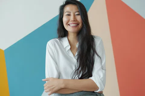 Smiling woman with long dark hair wearing a white shirt standing against a colorful geometric wall.