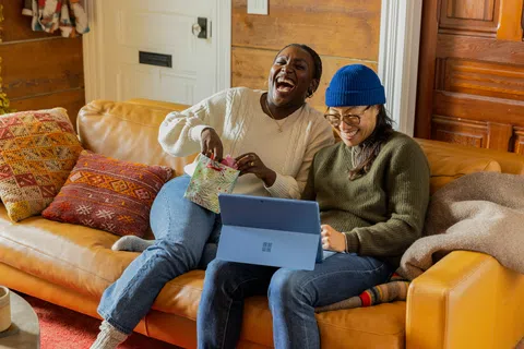 Two friends laughing and using a laptop while sitting on a cozy orange couch with colorful pillows.