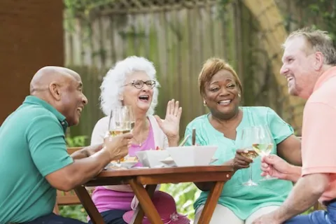 Four senior friends enjoying outdoor meal with white wine glasses around wooden table in garden.