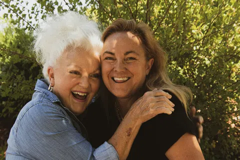Two smiling women, one elderly with white hair and one middle-aged, embracing in a garden outdoors.