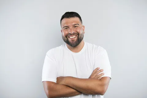 Smiling man with crossed arms wearing a white t-shirt against a plain light gray background.
