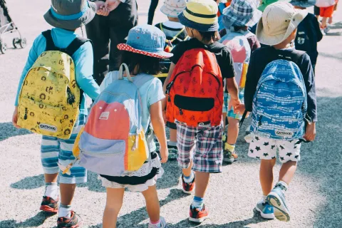 Group of young children wearing colorful backpacks and hats walking outdoors in a sunny setting.