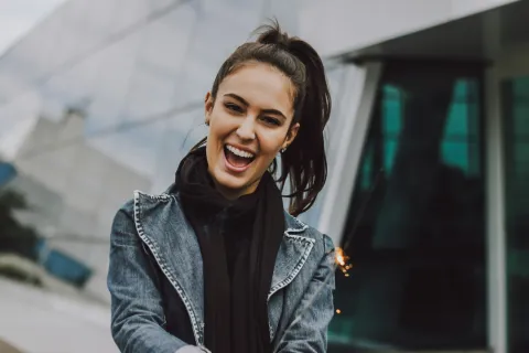 Smiling woman in denim jacket and scarf, standing outdoors with modern architecture in the background.