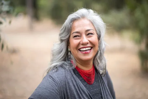 Smiling woman with silver hair and red necklace in a natural setting, exuding warmth and positivity.