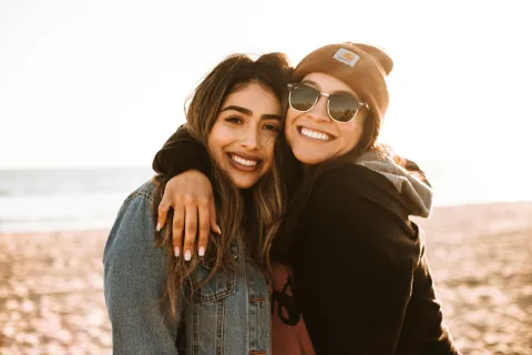 Two young women smiling and hugging at the beach during sunset, one wearing sunglasses and a beanie.