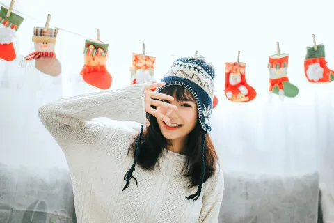 Young woman in cozy sweater and winter hat sitting on couch with Christmas stockings hanging behind her.