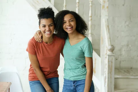 Two young women in casual clothes smiling and hugging indoors with a white rustic staircase background