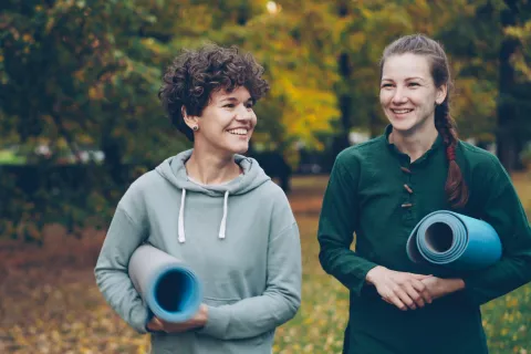 Two women smiling and holding yoga mats walking outdoors in autumn with fallen leaves and trees.