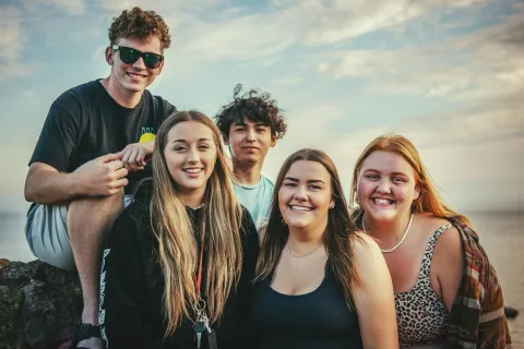 Group of five smiling young adults posing outdoors with a scenic sky background during sunset.