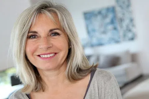 Smiling middle-aged woman with blonde hair in a casual top in a bright, modern living room setting.